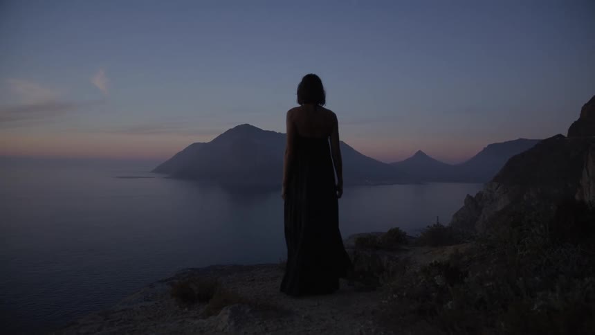 View from behind of a woman wearing a long dress walking on a cliff overlooking the sea at dawn in shades of blue and purple