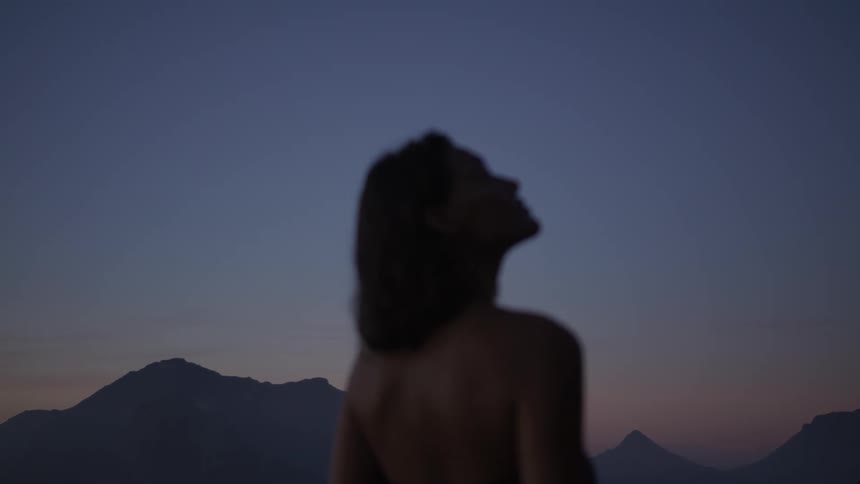 A woman stands in front of a mountain range looking up at the night sky