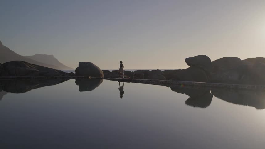 Woman walking next to rocks and a lake with the silhouette of mountains on the horizon on a morning