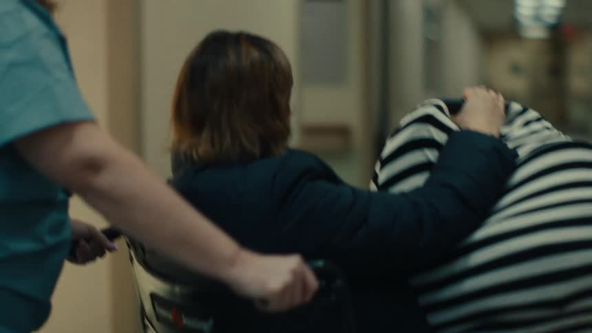 Woman being carried in a wheelchair by a nurse in a hospital hallway
