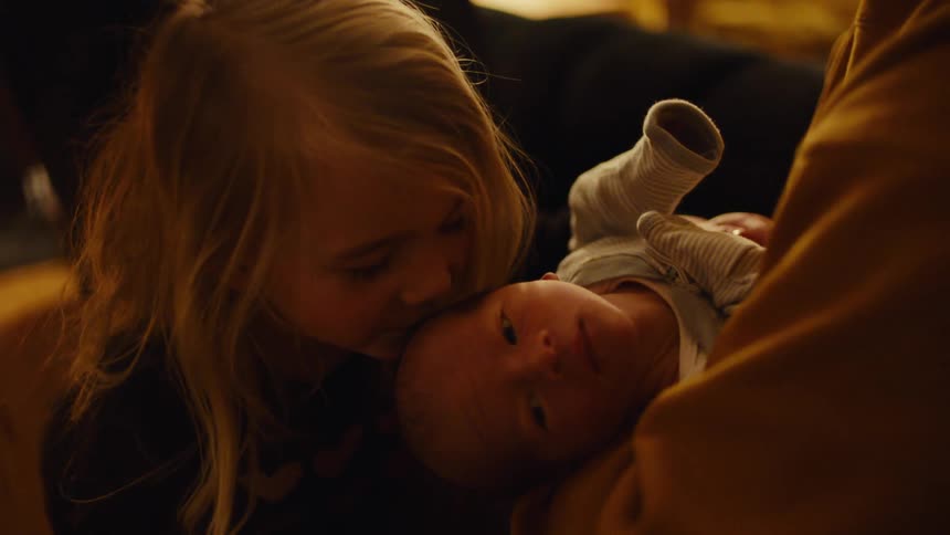 A little blonde girl kissing the head of a newborn baby who is on an adults lap