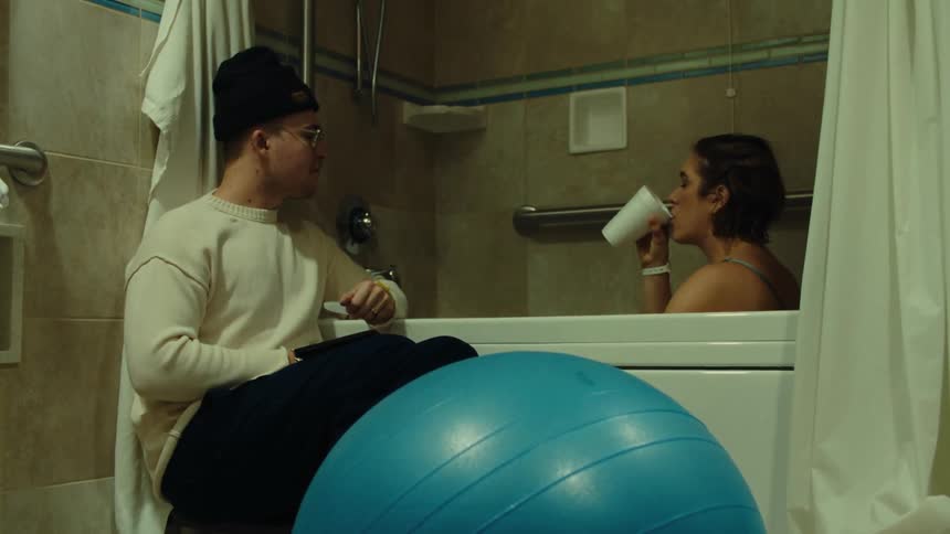 A girl sits in the tub and drinks from a cup while the guy sits next to the tub