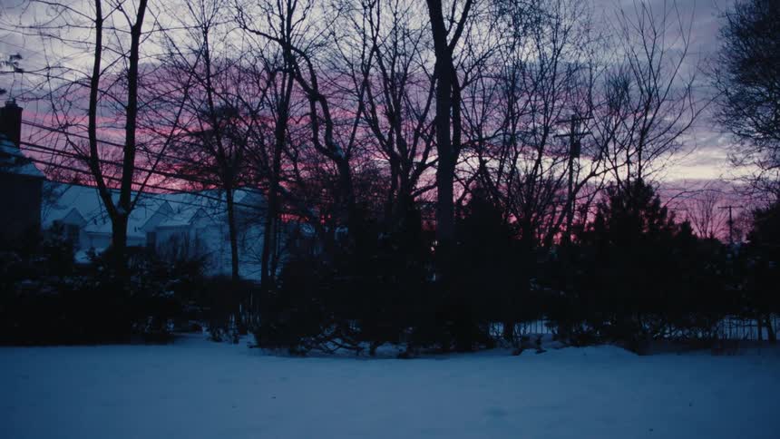 Leafless trees silhouetted against a red sky separate a large house from a snow covered field