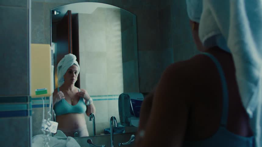 Woman with a towel on her head looking at herself in the mirror wearing only a bra