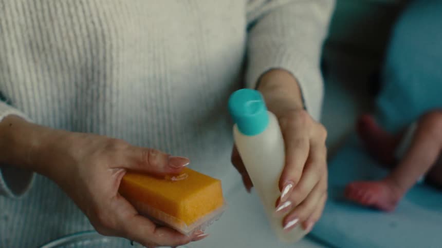 Hand putting liquid soap on a small sponge and the legs of a newborn baby in the background