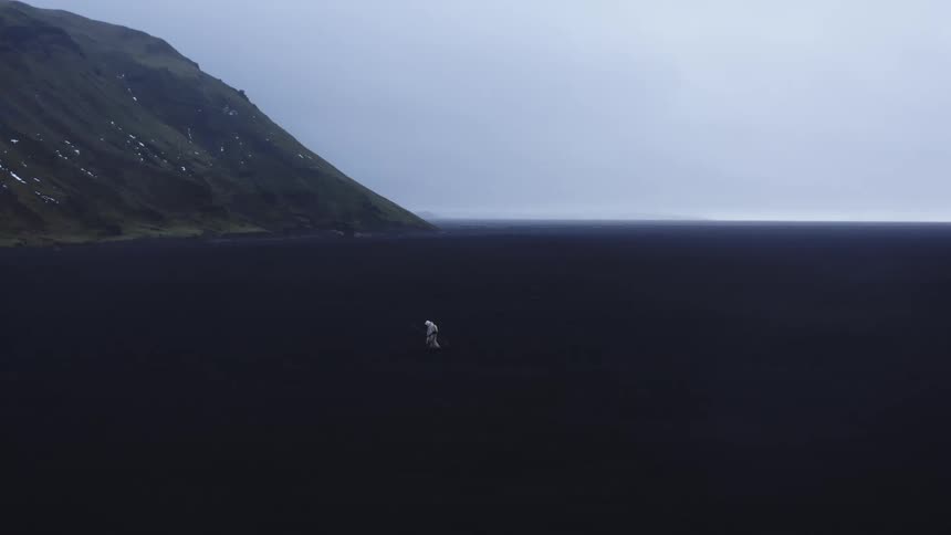 A person dressed all in white walking fast on a plain with dark terrain near a mountain