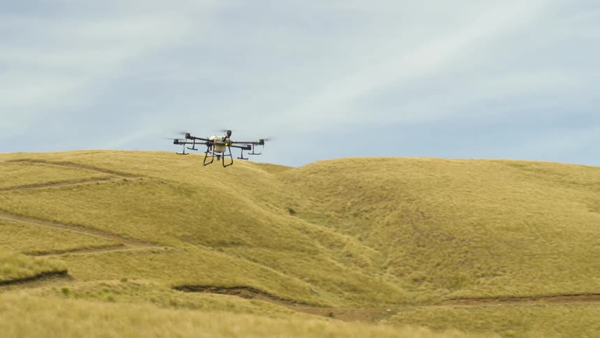 A large agricultural drone approaching to land on a grassy field with many hills