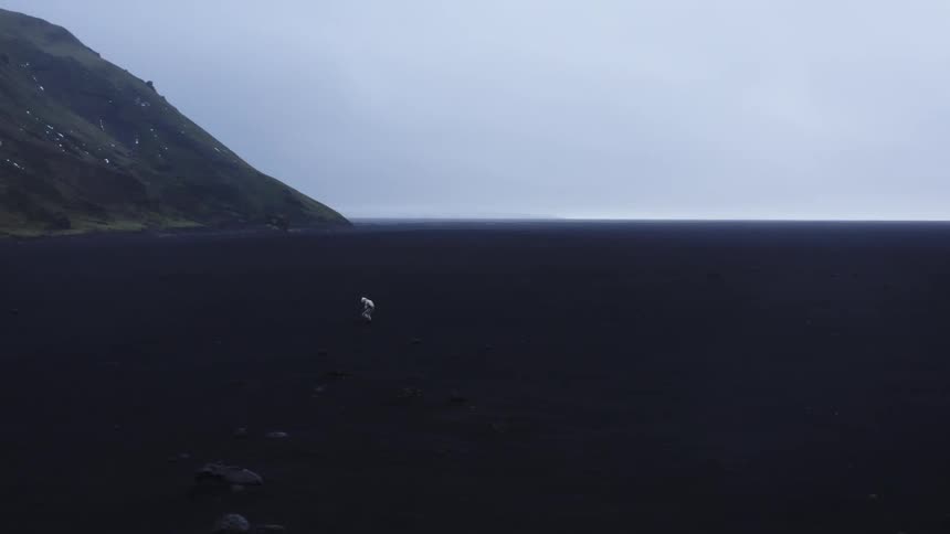 A figure in white walks across a vast black plain near a mountain