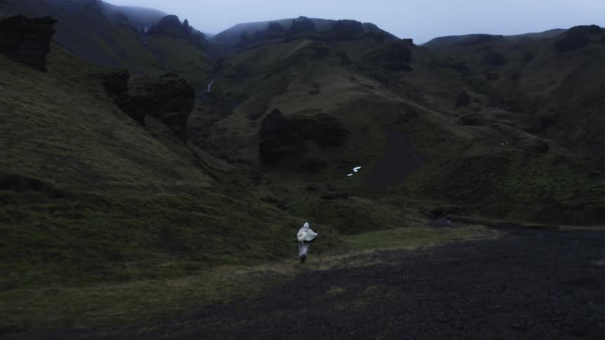 Person carrying a staff walks on a field along the base of a mountain