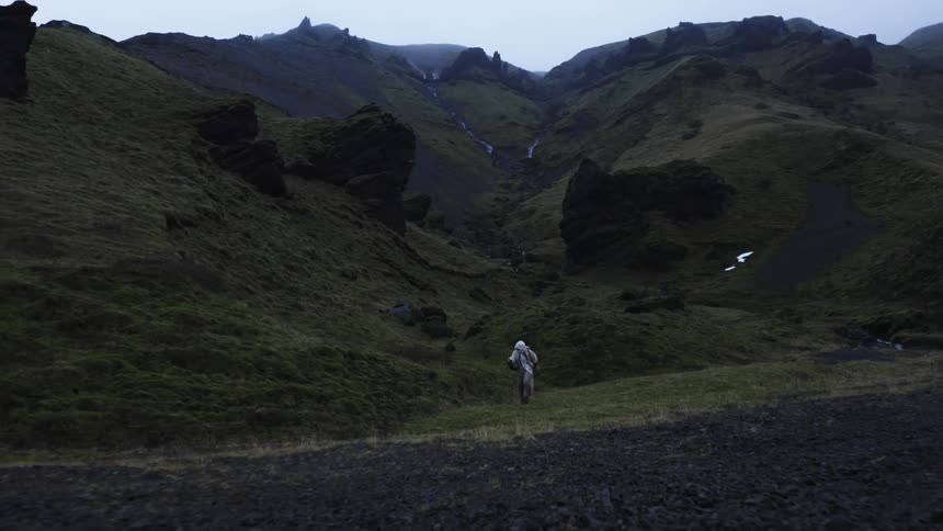 Man wearing loose clothing and carrying a staff walks off a hilly path and onto a gravel covered field