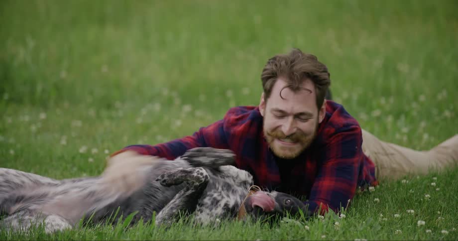 A man smiling as he pets a black and white dog in a field with both of them lying in the grass