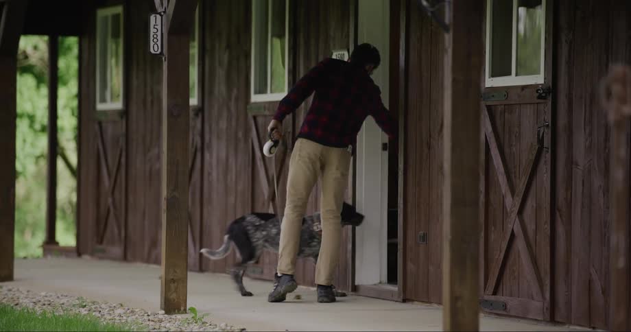 A man walking a dog alongside a wooden cabin entering the building through a door