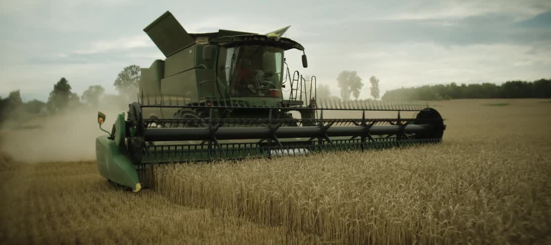 A green combine harvester operates in a golden wheat field under a cloudy sky