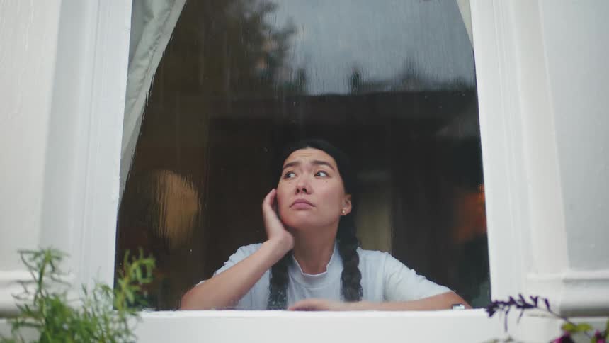 Smiling woman opens curtains only to frown and stare out the window at the rain