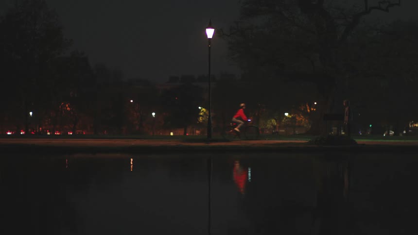 Person walking and another riding a bike in a park next to a lake at night