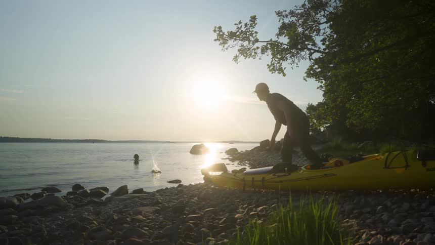 A person stands near a yellow kayak on a rocky shoreline with a woman bathing on a lake extending into the distance