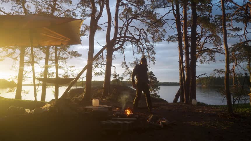 Man by a lake in a natural setting next to a campfire at a campsite
