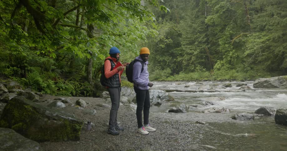 Two black hikers stop at a river so one of them can remove a book from his friends backpack