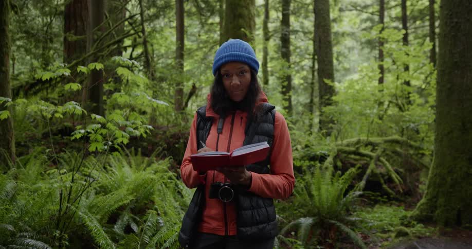 A woman standing in the forest writing on a notebook looks at the camera