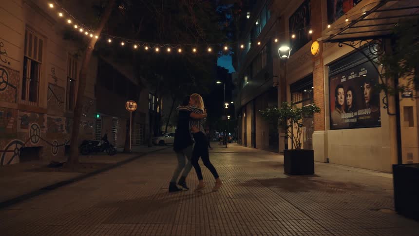 Couple dances arm in arm down an empty street passing shops at night