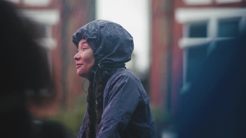 A woman wearing a raincoat with a hood stands outdoors while raining