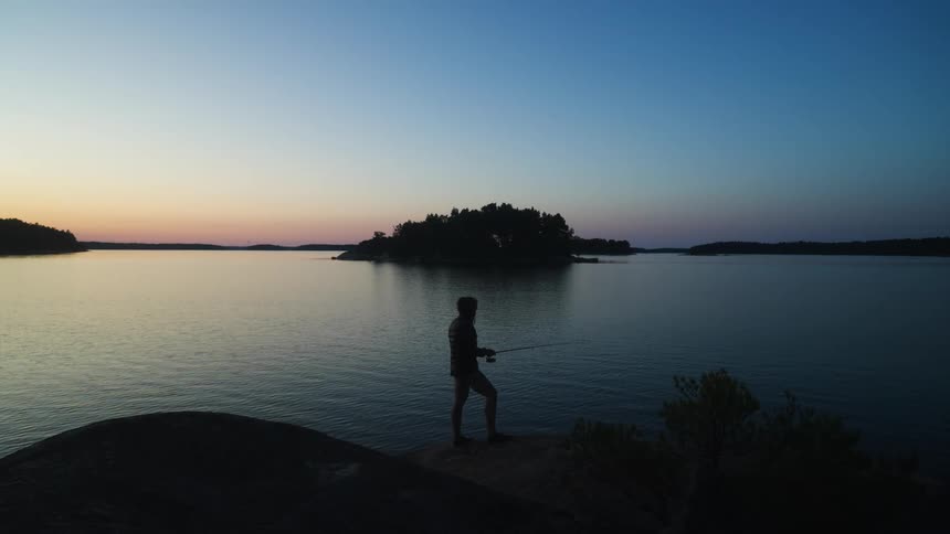 Man fishes in a lake as red tints the horizon and casts him partially into a silhouette