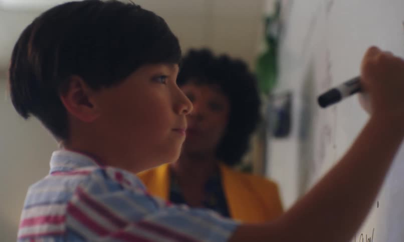 A little boy is writing something on a whiteboard using a black pen and then he gives a smile