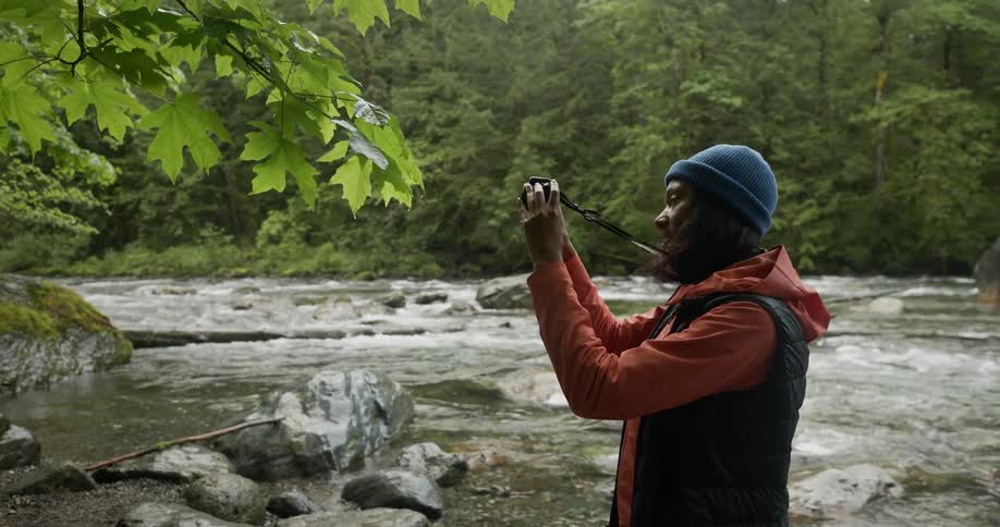 A woman holding a camera takes pictures of plants growing near a river in the forest