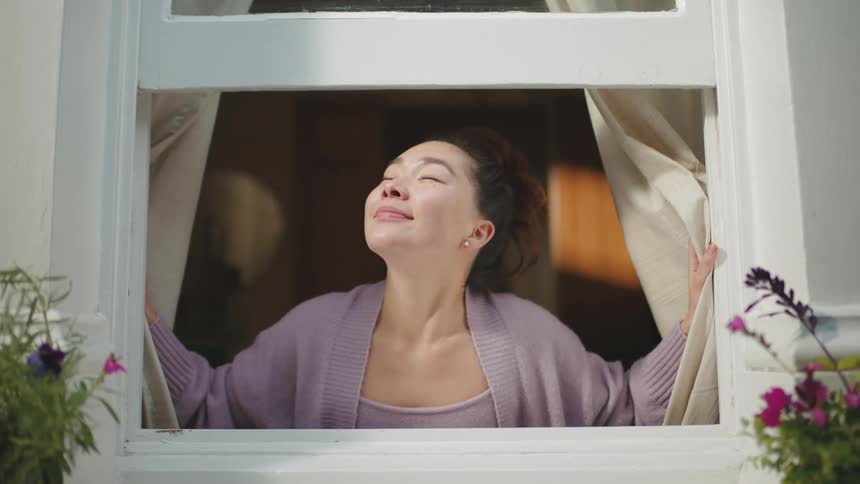 Woman opening curtain from an open window and breathing in the air outside