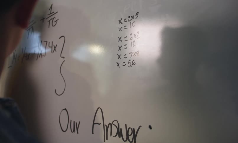 A teenage student works out a problem on a whiteboard as his teacher looks on