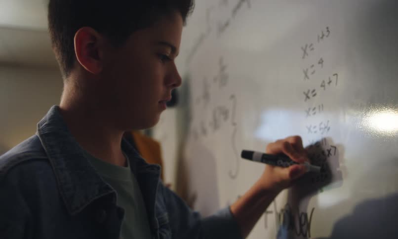 A student writes mathematical equations on a whiteboard next to a teacher