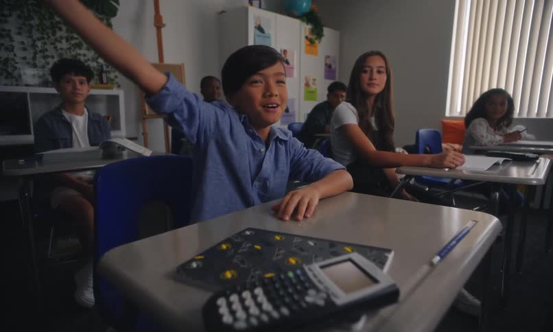 A classroom scene with students where a boy is raising his hand to answer a question while others are seated at their desks