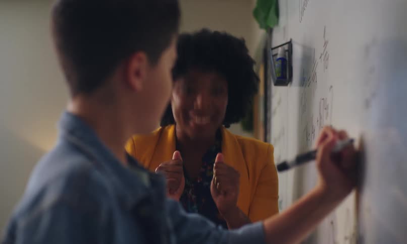 Boy writing on a whiteboard with the teacher next to him