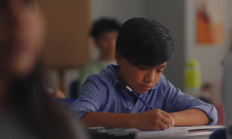 A student in a blue shirt sits at a desk in a classroom focused on writing or working on an assignment