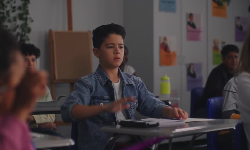 A student seated at his desk in school raises his hand and walks toward the front of the classroom