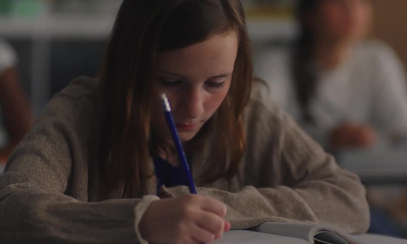 A girl seated at a desk writing in a notebook with a pen at school