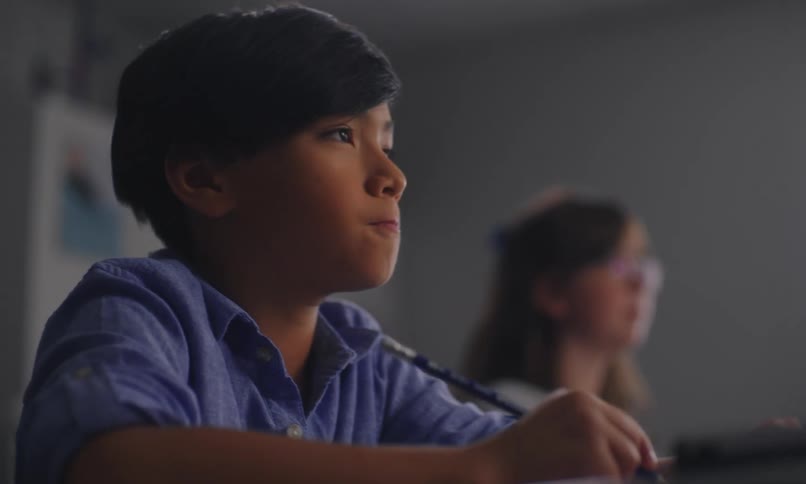 Boy sitting in the classroom writing and a girl classmate in the background