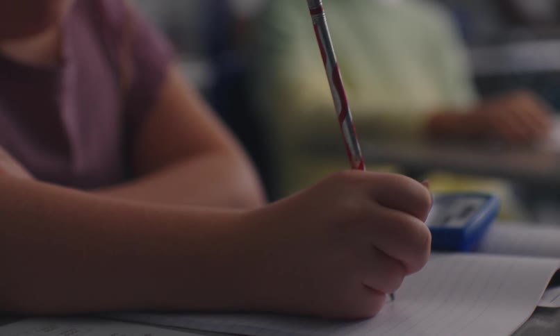 A close up of a childs hand holding a pencil while writing on a notebook in a classroom setting
