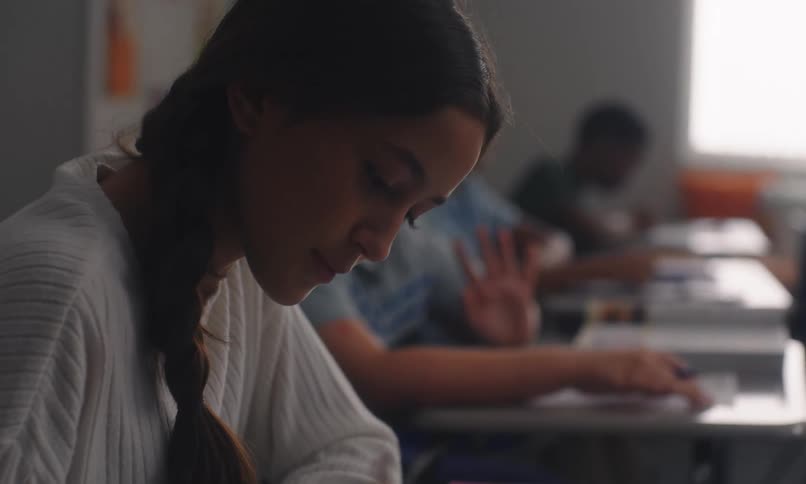 A young woman focused on writing in a classroom with several classmates visible in the background