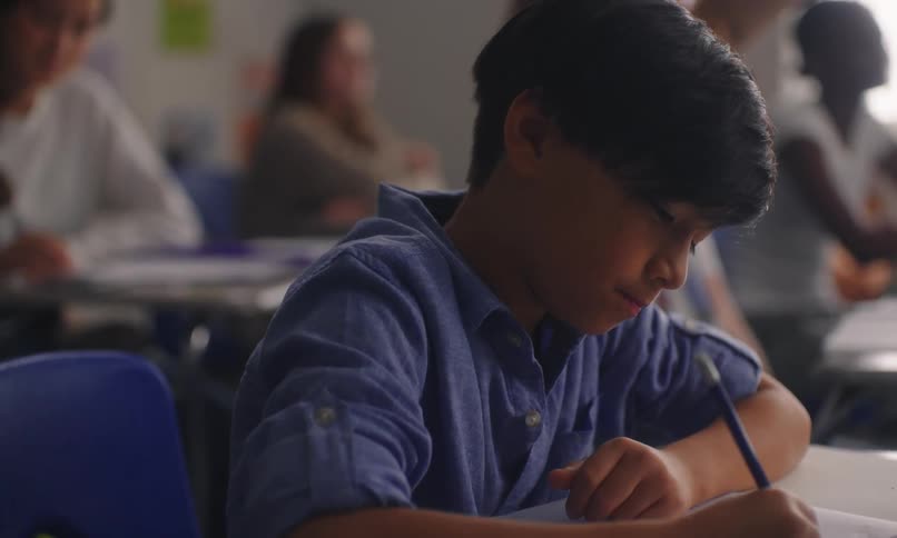 Boy sitting in a classroom writing with other students in the background