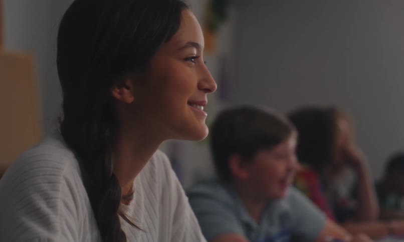 A young girl is writing something on a paper using a pencil and she looks forward and smiles