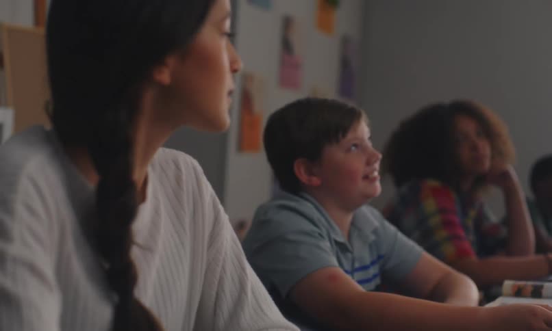 A young girl raising her hand in a classroom setting with other students visible in the background