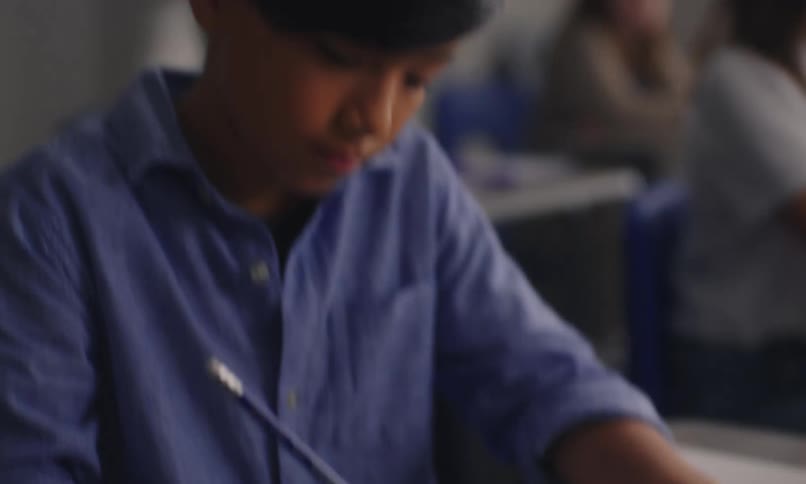 A student in a classroom sits at a desk and writes in his notebook