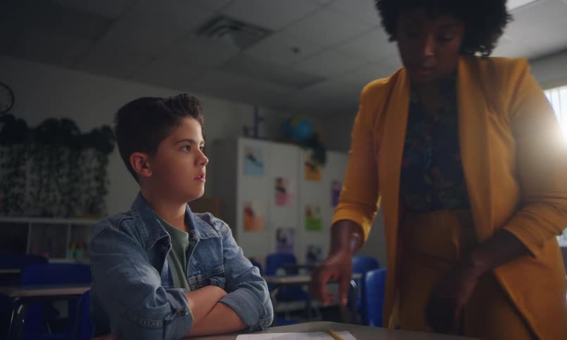 A little boy is writing something on a paper in a school room and then a woman looks at the paper to correct it