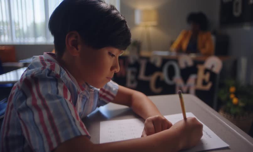 Student works on an exam while his teacher sits at her desk and watches