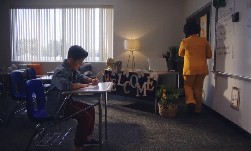 A teacher in a yellow outfit engaging in a conversation with a student seated at a desk in a bright classroom setting