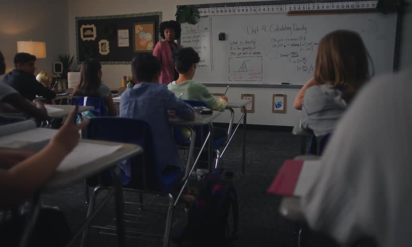 A teacher walks back and forth at the front of the classroom and calls on students raising their hands