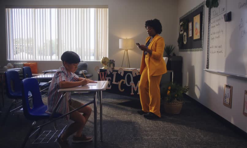 A classroom with a student sitting at a desk focused on his work while a teacher stands nearby checking her phone
