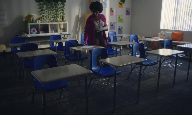 A teacher places tests inside an orderly classroom with blue chairs and desks