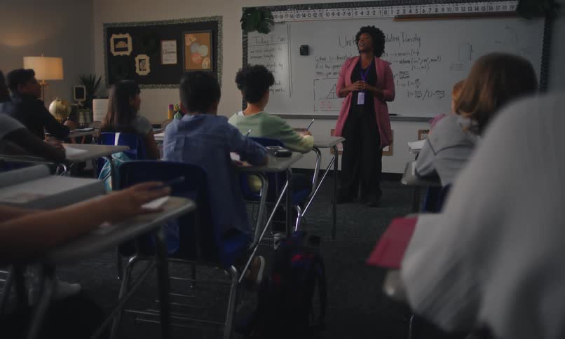 A classroom with students seated at desks facing a whiteboard where a teacher gestures during a lecture